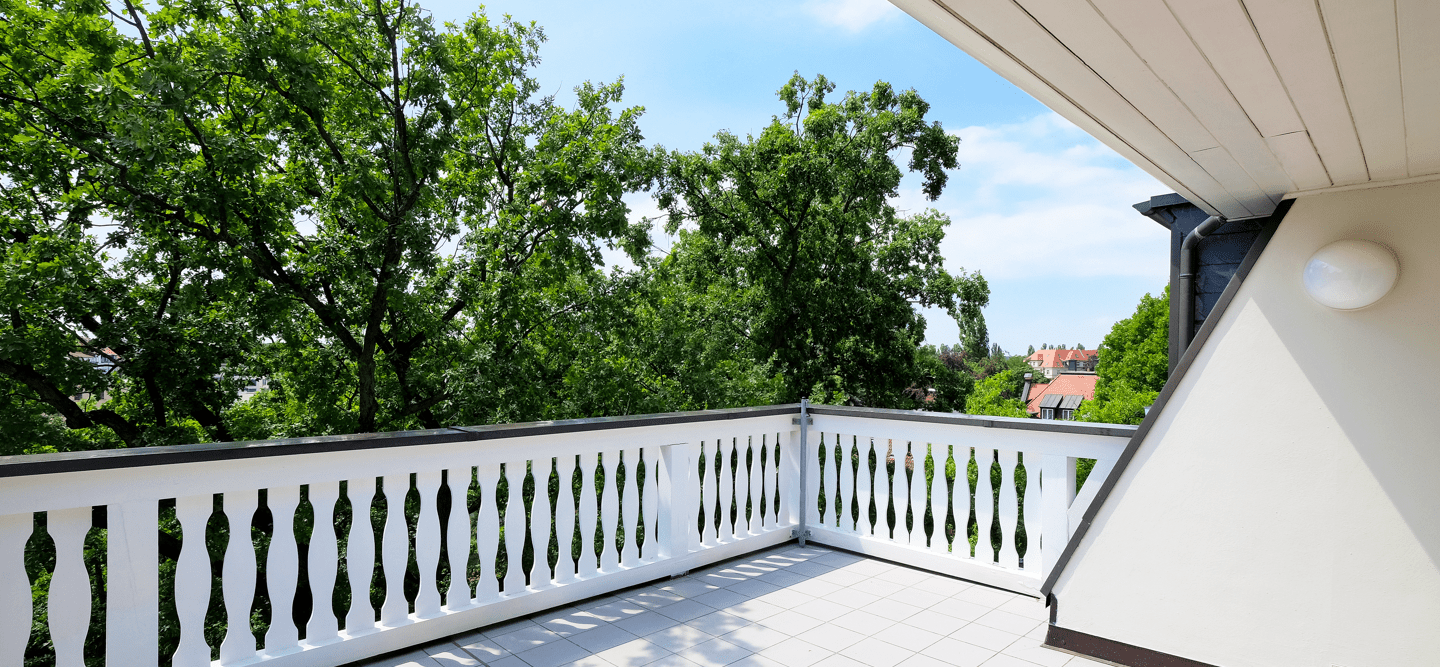 Ein Foto einer Dachterrasse mit Blick auf grüne Baumkronen und blauen Himmel. Der Boden ist gefliest und das Geländer besteht aus weißem Holz.