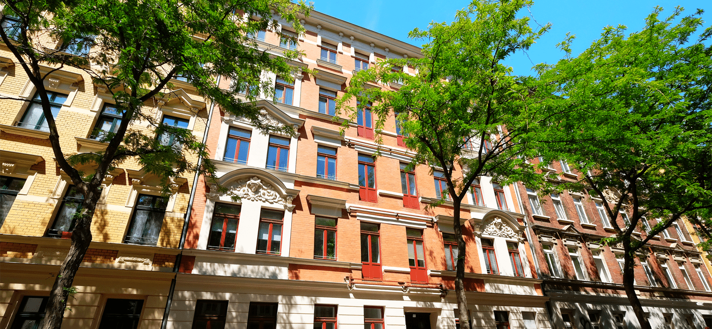 Ein Foto einer sanierten Altbau-Fassade. Das Mauwerk besteht aus beigen und roten Backsteinen. Davor stehen zwei grüne Bäume.