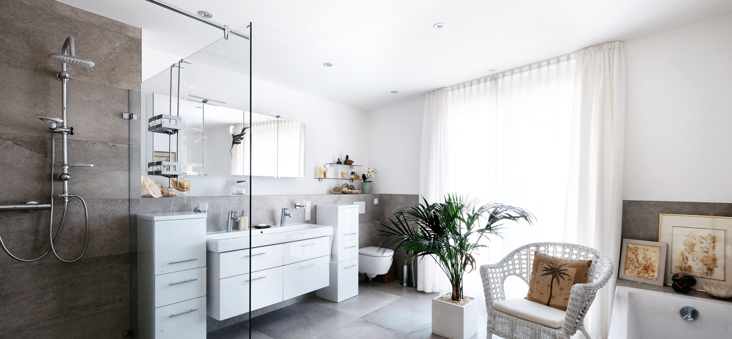 Ein Foto eines geräumigen, hellen Bads mit Wanne, Dusche, Waschbecken sowie einer Palme und diversen Dekoartikeln. Die Fliesen sind grau, die Wände weiß. Ein Fenster erhellt den Raum.