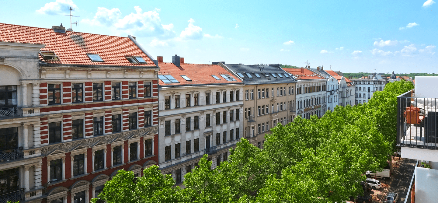 Foto vom Dachgeschoss aus auf gegenüberliegende Dächer und Fassaden von Denkmalimmobilien. Weiter unten sind grüne Baumkronen zu sehen.