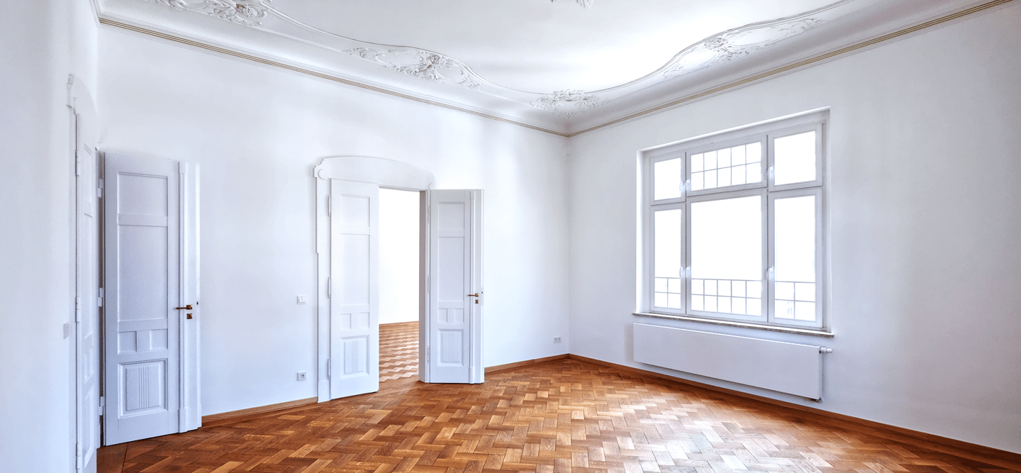 Foto eines großen Raums mit weißen Wänden, Stuck, Echtholzparkett und zwei historischen Flügeltüren sowie einem großen Fenster.