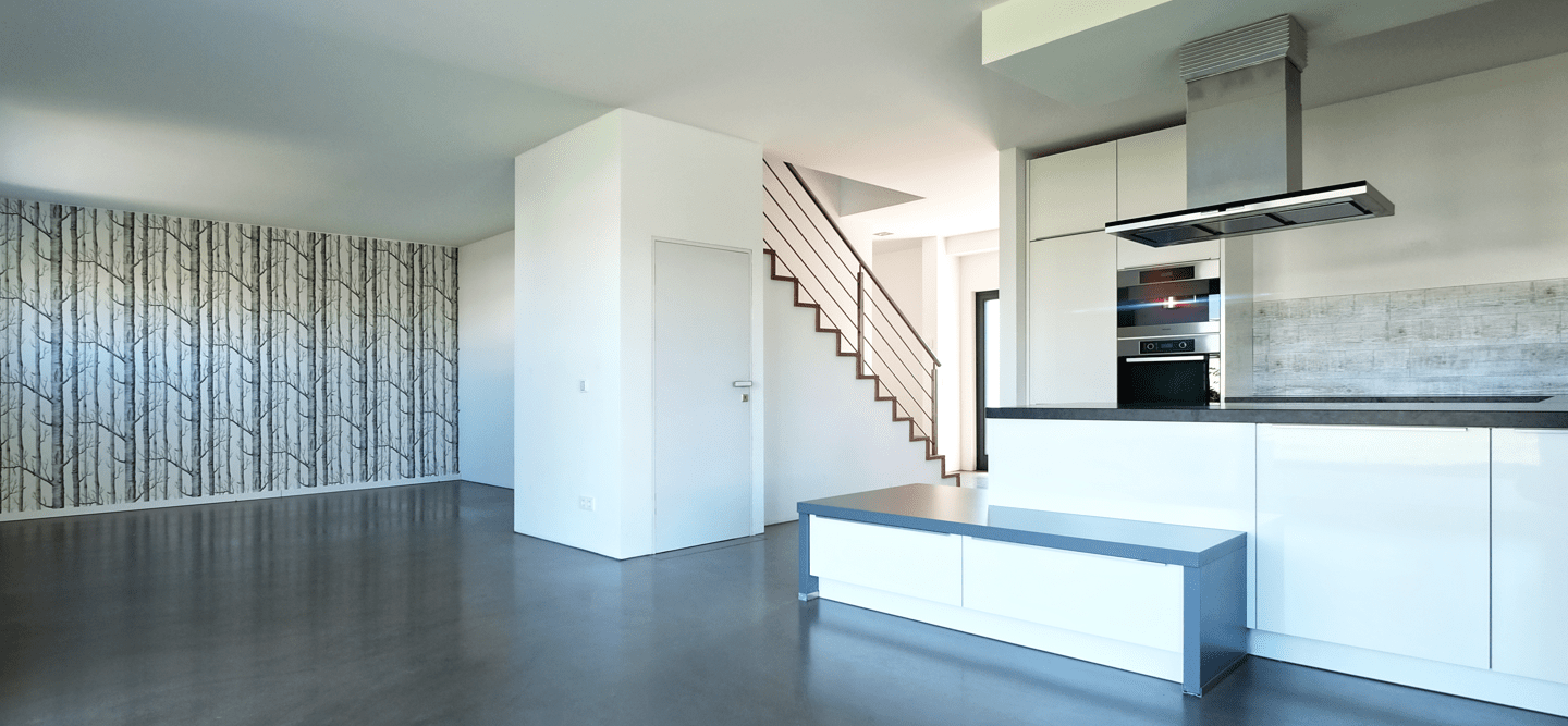 Foto eines Wohnzimmers mit offener, heller Küche, Treppe zum Obergeschoss, einer tapezierten Wand und grauem Boden.