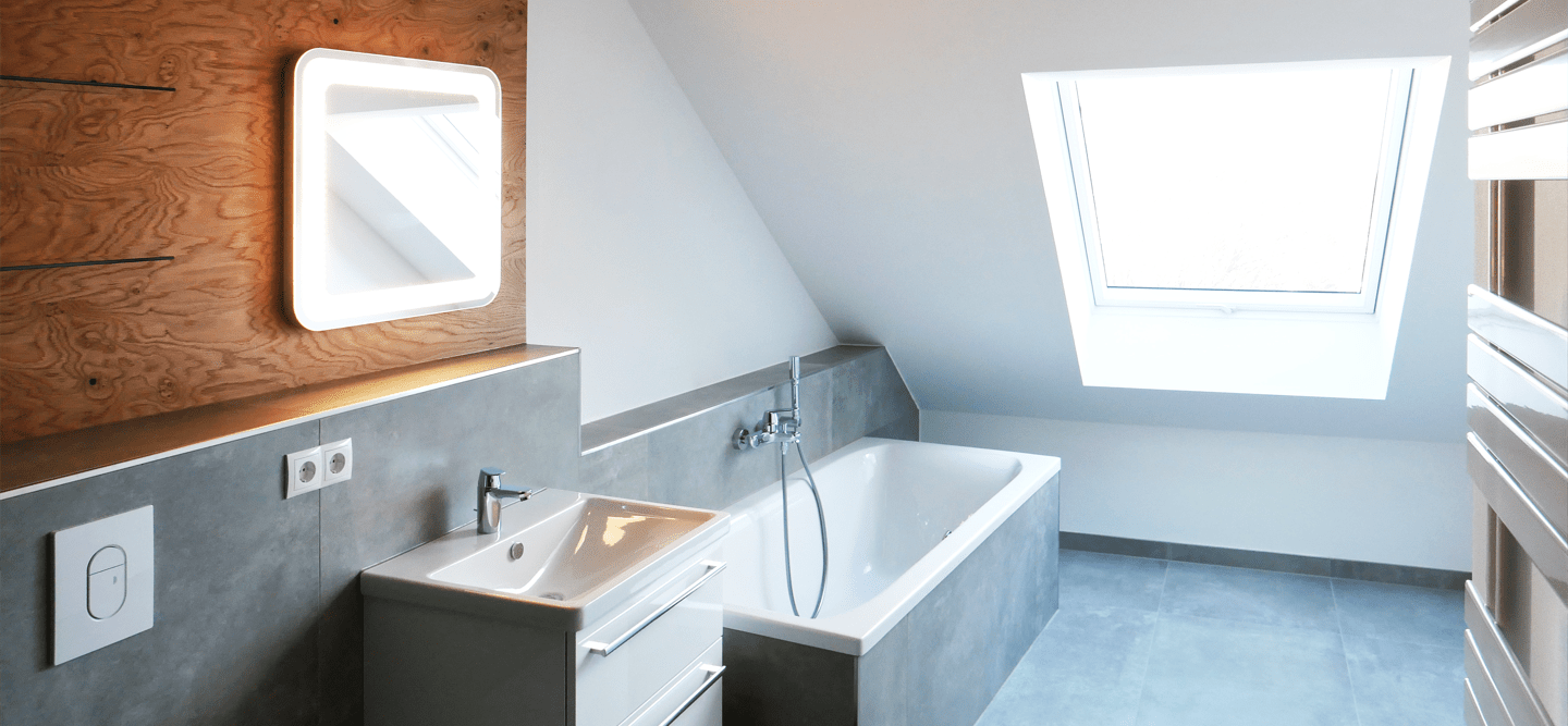 Foto eines modernen Bads mit Fenster im Dachgeschoss. Man sieht eine Wanne, einen Waschschrank, einen beleuchteten Spiegel, Holzverkleidung, graue Fliesen und einen Handtuchheizkörper.