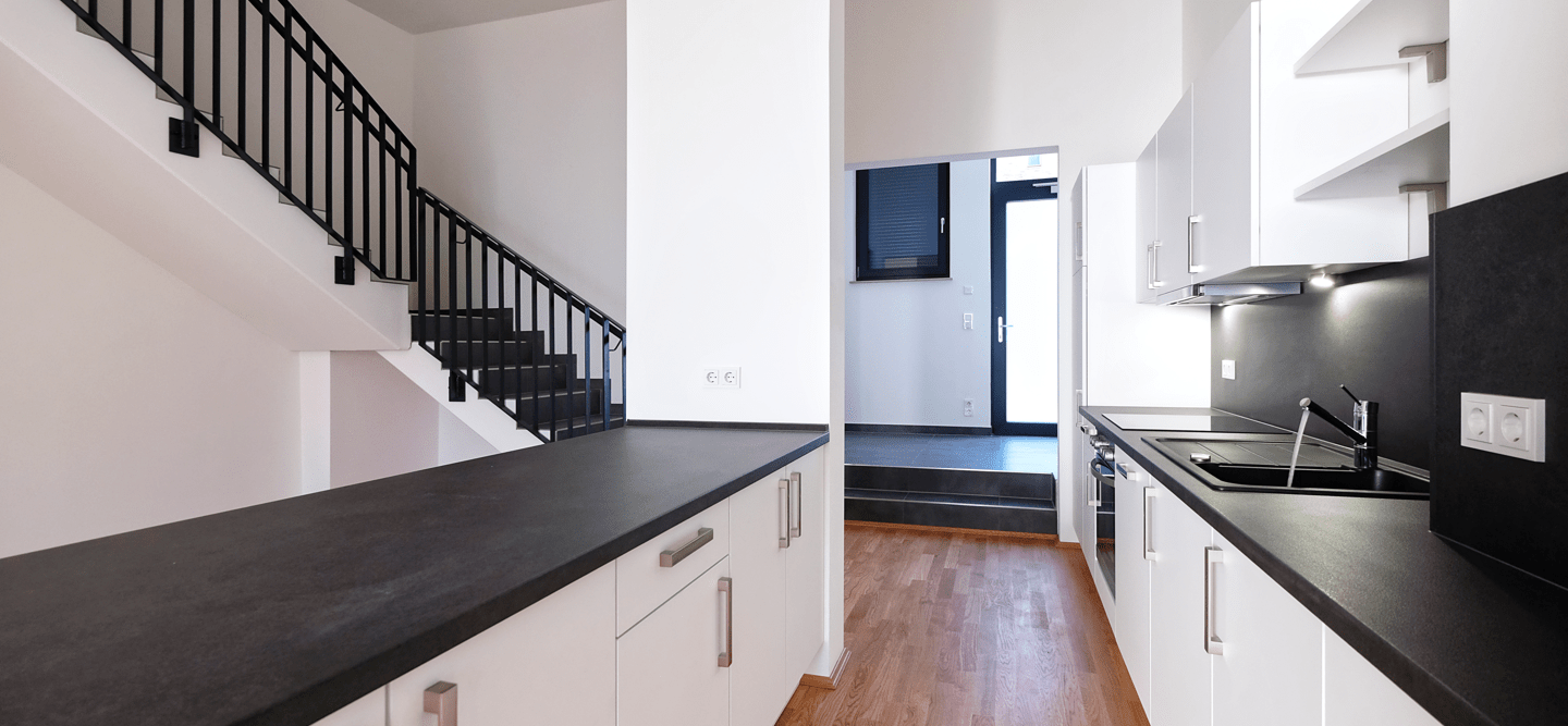 Foto einer offenen Küche mit Kochinsel, Küchenzeile, Zugang zur Wohnungstür sowie Eck-Treppe in ein Obergeschoss.