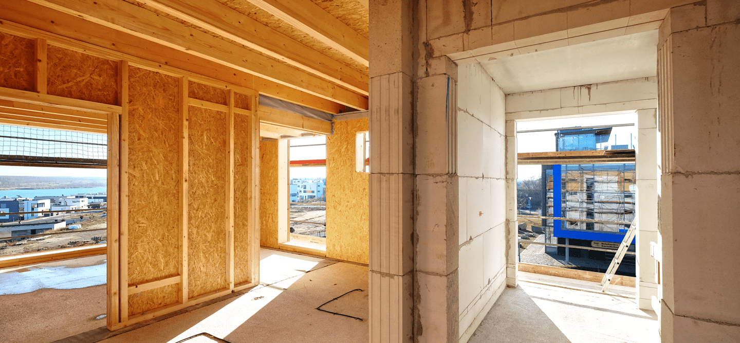 Foto einer Wohnung im Rohbau. Man Sieht Betonwände, Holzwände und Holzdecken. Durch die Aussparungen für Fenster und Türen scheint die Sonne herein.