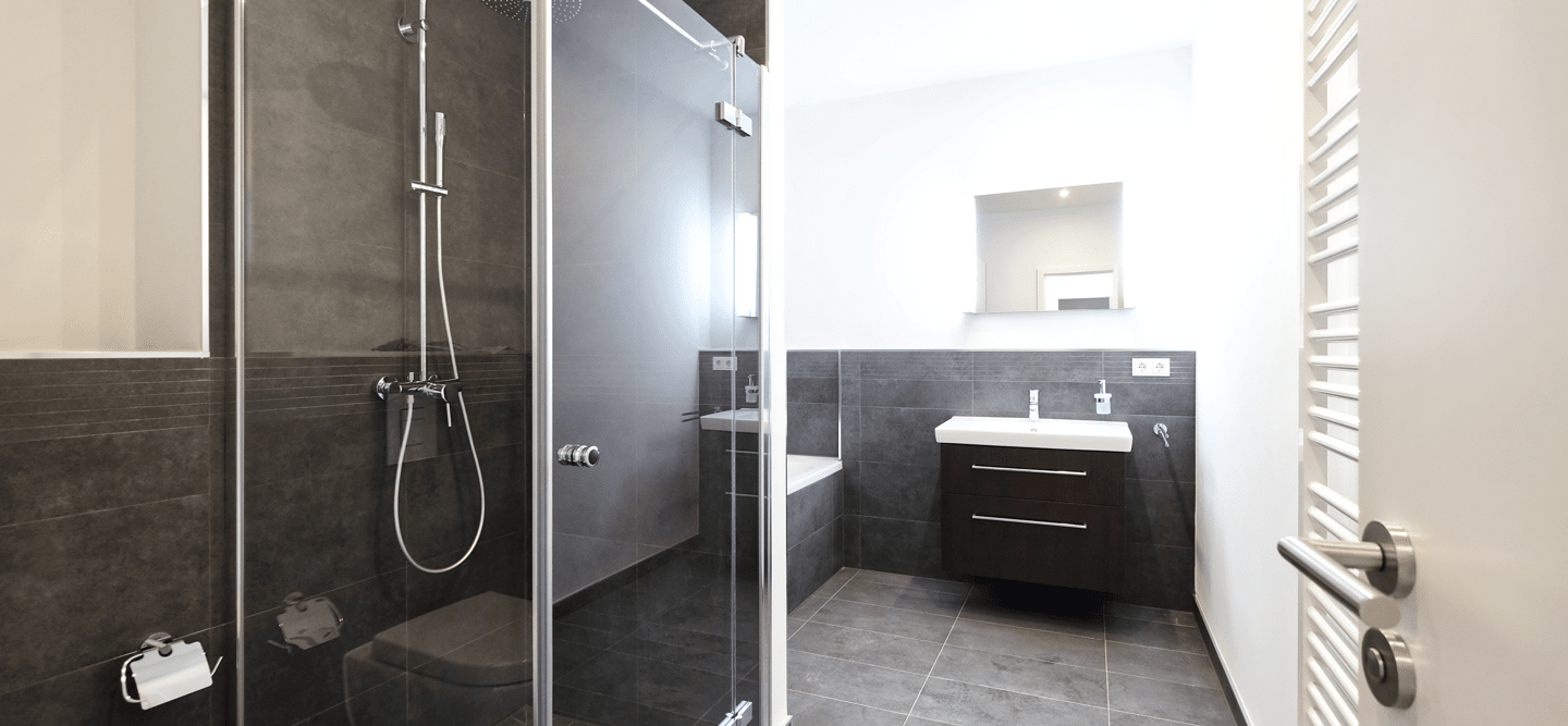 Foto eines innenliegenden Neubau-Bad mit Dusche, Wanne, Spiegel und Waschbecken mit Unterschrank. Die Fliesen sind dunkelgrau, die Wände weiß.