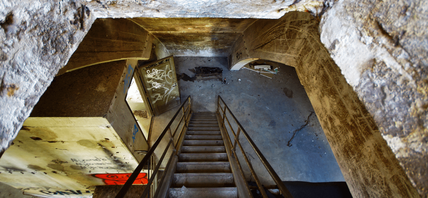 Foto mit Blick von einer Treppe, welche nach unten in einen Raum führt. Alles ist baufällig.