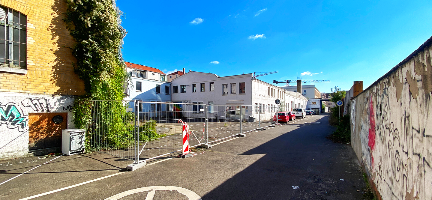 Foto einer leerstehenden, alten Fabrikhalle in einer Seitenstraße. Gegenüber verläuft eine Mauer. Hier entsteht das Wohncarré Plagwitz.