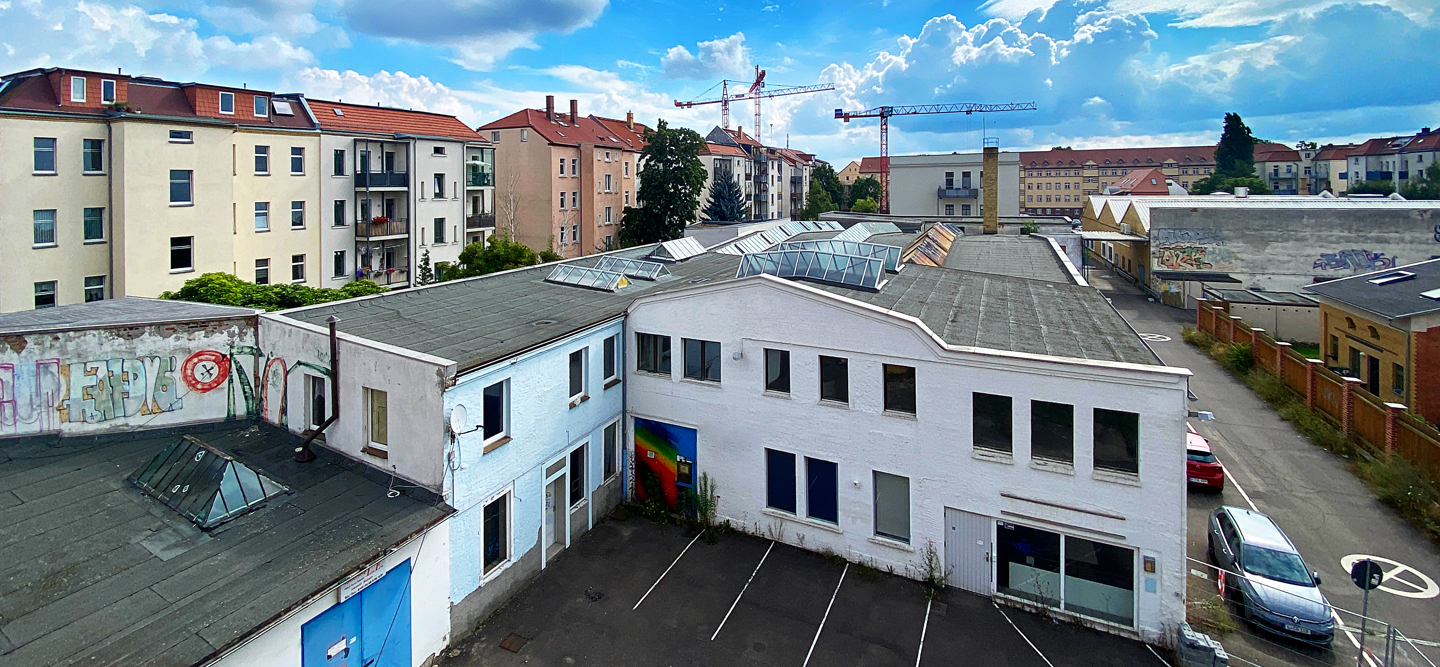 Luftbildaufnahme einer kleinen Straße mit alten Industribauten und sanierten Immobilien im Hintergrund. Der Himmel ist blau und bewölkt. Hier entsteht das Wohncarré Plagwitz.